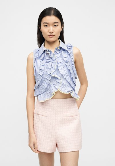 Νεαρή γυναίκα που φοράει μια μακρυμάνικη μπλε ριγωτή κοντή μπλούζα και ροζ υφασμάτινα σορτς με ψηλή μέση, στέκεται μπροστά σε απλό φόντο.