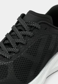 Chaussure de sport noire avec des lacets blancs à pois et un motif hexagonal texturé sur la partie supérieure, reposant sur une semelle blanche.