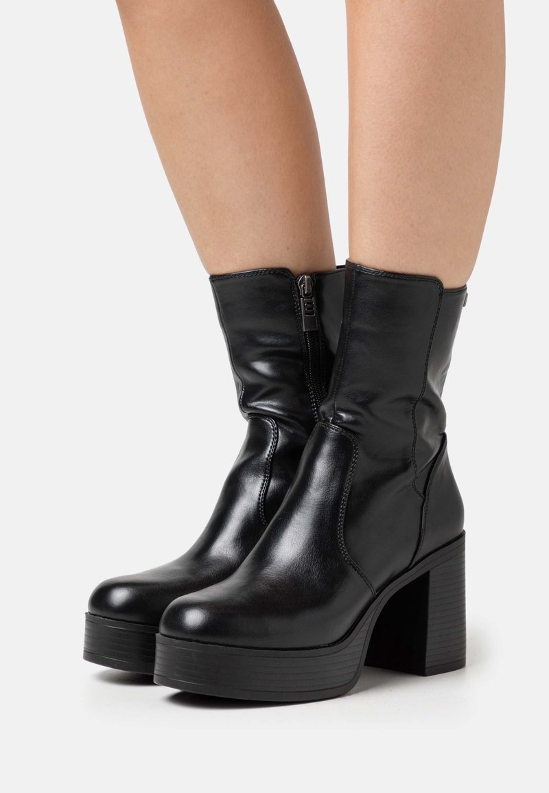 Bottes plates-formes en cuir noir mi-mollet avec talons épais et fermetures éclair latérales, portées sur des jambes nues sur fond blanc.