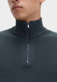 Dunkelgrüner, gerippter Strickpullover mit hohem Kragen und halbem Reißverschluss. Verfügt über eine feine Textur und minimalistisches Design.