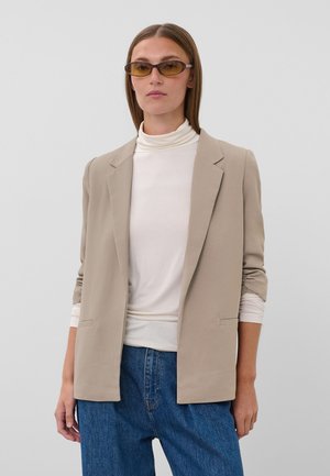 Femme portant un blazer beige, un col roulé blanc, un jean bleu et des lunettes de soleil teintées ambre, debout devant un fond gris clair uni.