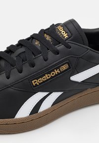 Svart lädersneaker med vita detaljer, texturerad gummisula och logotyp i guld. Har svarta snören och klassiska designlinjer.