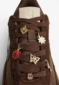 Brązowe zamszowe sneakersy z złotymi zawieszkami na sznurówkach: serce, kwiat, motyl oraz czerwona kolczasta owoc. Prosty kremowy design języka.