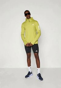 Roheline pikkade varrukatega sporditopp, milles on pooltõmbluku detail, valged aktsendid ja kolm triipu varrukatel, koos mustade lühikeste pükste ja tumesiniste spordijalatsitega.