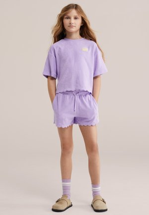 Junges Mädchen steht, trägt ein passendes lila Kurzarmpullover- und Shorts-Set mit gezackten Kanten und helllila gestreiften Socken.