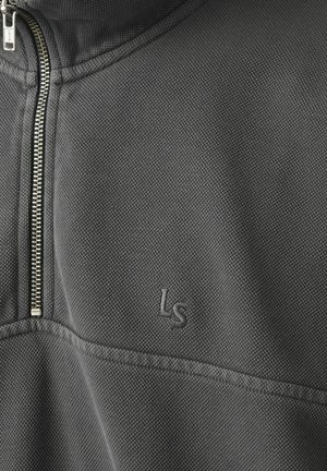 Lähikuva tummanharmaasta teksturoidusta kankaasta, jossa on hopeanvärinen vetoketju ja ommeltu "Ls"-logo vaatteen rintakehän alueella.