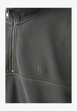 Nahaufnahme eines dunkelgrauen, strukturierten Stoffs mit einem silbernen Reißverschluss und einem auf der Brustbereich eines Kleidungsstücks aufgestickten "Ls"-Logo.