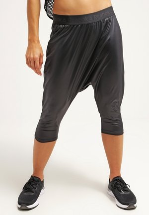 Person, die schwarze Reebok-Caprihosen mit tiefem Schritt und schwarze Sportschuhe trägt, steht mit gespreizten Beinen vor weißem Hintergrund.