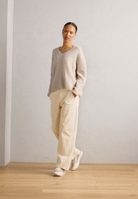Maglione di lana beige chiaro con scollo a V, abbinato a pantaloni larghi crema e sneaker chiare, su uno sfondo neutro.
