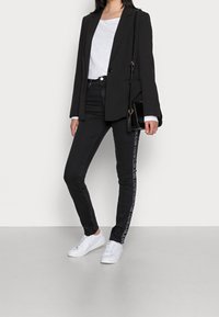 Blazer negro sobre una camiseta blanca, combinado con jeans negros con una marca blanca a lo largo del costado y zapatillas blancas. Texturas suaves.