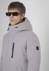Lichtpaarse gewatteerde jas met een hoge kraag en een verstelbare capuchon. Bevat zwarte ritsen en een logopatch op de linker schouder.