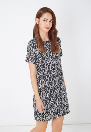 Joven mujer con cabello largo castaño está vestida con un vestido negro con un patrón de bucle abstracto en blanco, frente a un fondo blanco liso.