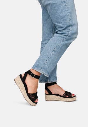 Sorte espadrille-kilesandaler med flettet design, ankelrem og jute-platform. Båret med lyseblå, korte jeans.