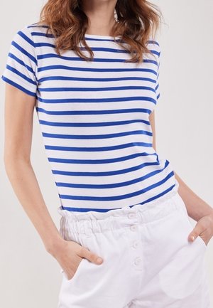 Mujer con una camiseta de manga corta a rayas azules y blancas metida en pantalones blancos de talle alto con botones, manos en los bolsillos.