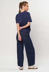 Ženska s kitkami, oblečena v temnomodro majico, temne široke kavbojke, bele superge in obroče za ušesa, stoji z rokami za hrbtom.