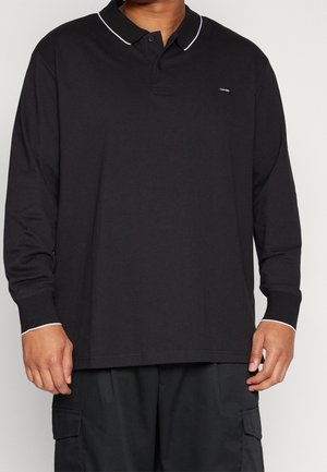 Muž oblečený v černém polo tričku s dlouhým rukávem a bílým lemem na límci a manžetách, doplněném černými vojenskými kalhotami.