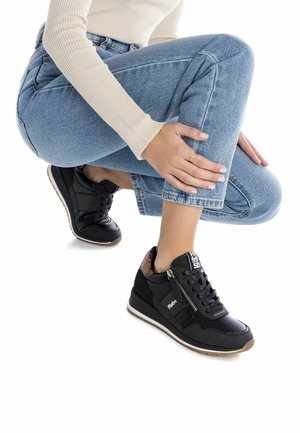 Zapatillas deportivas negras con acabado brillante, cuello acolchado, detalle de cremallera lateral y parte trasera texturizada en contraste. Se presentan sobre unos vaqueros claros.