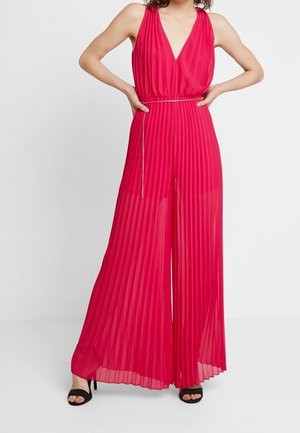 Lyserød plisseret jumpsuit med dyb V-hals, markeret talje og et tilhørende metallisk bælte. Bukserne er vidde med transparent, flydende stof.