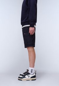 Vista laterale di una persona che indossa pantaloncini cargo blu navy, felpa blu navy, calzini bianchi con scritte nere e scarpe da ginnastica nere e bianche.