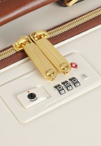 Maleta rígida con una superficie de color blanco cremoso, tiradores de cremallera dorados, un candado TSA y un dial de combinación con botones numéricos negros.