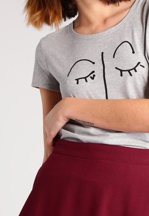 Vrouw die een grijs T-shirt draagt met een eenvoudig zwart gezicht ontwerp en een bordeauxrode rok, met een kleine tatoeage zichtbaar op haar onderarm.