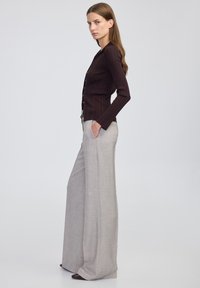 Frau, die seitlich steht, trägt einen dunklen, gerippten, geknöpften Pullover und weite, hellgraue Hosen mit den Händen in den Taschen.