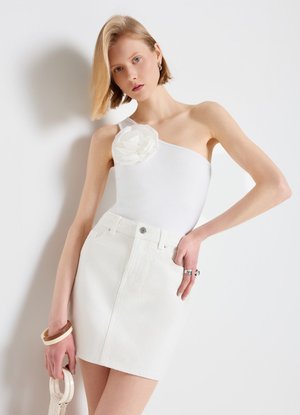 Giovane donna con capelli biondi corti che indossa un top bianco monospalla con dettaglio floreale e gonna bianca, che tiene una borsa braccialetto bianca e marrone.