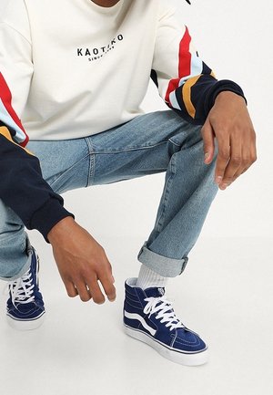 Modré vysoké tenisky s bílými podrážkami a tkaničkami, s texturovaným plátěným materiálem a detaily švů. Model nosí světlé džíny a krémovou košili.