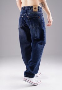Dark blue denim jeans featuring a relaxed fit, back pockets, and a leather patch on the waistband. Paired with white sneakers.