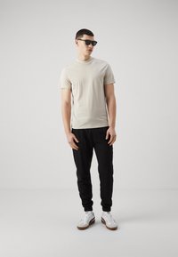 Camiseta beige de manga corta, joggers negros, zapatillas blancas con suelas marrones y gafas de sol negras. Diseño simple y colores neutros.