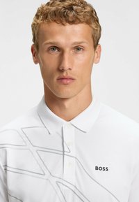 Weißes Poloshirt aus glattem Material mit einem grauen abstrakten Muster und einem schwarzen "BOSS"-Logo auf der Brust, klassischem Kragen und Knöpfen.