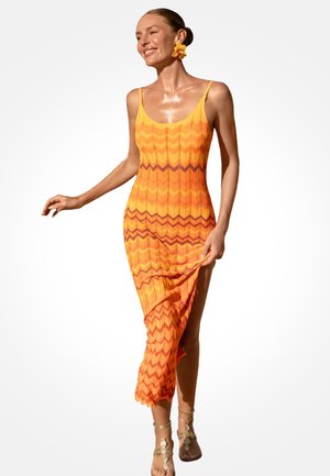 Femme en robe maxi sans manches à motif zigzag orange, tenant l'ourlet, souriante, portant une boucle d'oreille fleur et des sandales dorées, debout sur fond blanc.