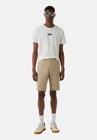 T-shirt blanche avec le logo "BOGNER", short beige, baskets blanches et bleu marine, et lunettes de soleil grises. Le modèle se tient devant un fond uni.