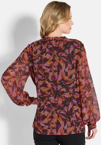 Transparente Bluse mit langen Ärmeln, die einen gerafften Ausschnitt und ein buntes, abstraktes Blumenmuster in Lila-, Orange- und Schwarztönen aufweist.