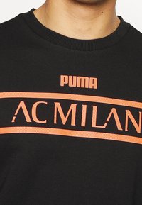 Felpa nera con la scritta "PUMA" e "AC MILAN" in testo arancione brillante. Collo a costine e texture liscia.