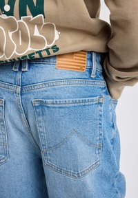 Jeans de mezclilla azul claro con corte recto, costuras en contraste y dos bolsillos traseros. Presenta un parche de cuero marrón con un diseño de bandera.