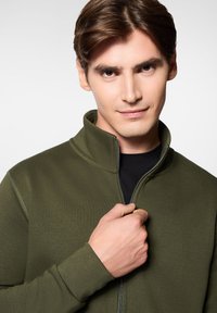 Oliwkowa polarowa kurtka zapinana na zamek z wysokim kołnierzem i ściągaczami na rękawach. Model trzyma zamek jedną ręką, czarna koszulka widoczna pod spodem.