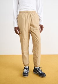Beige letvægt tapered bukser med elastisk talje og sidelommer, parret med sorte sportssko med en hvid snøredesign.