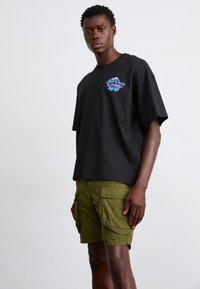 Black cotton t-shirt with a multicolored graphic logo on the chest, paired with olive green cargo shorts featuring multiple pockets.