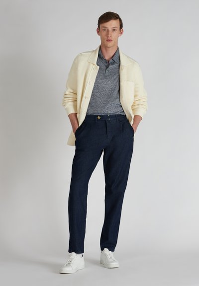 Crèmekleurige gebreide cardigan over een grijze polo, gecombineerd met donkerblauwe broeken en witte sneakers. Opvallende kenmerken zijn de textuur van de stof en een zak.