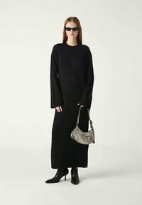 Sieviete, kura valkā garu melnu kleitu, melnus augstus papēžus ar norādītu galu, melnus saulesbrilles un tur mazu pelēku rokassomu.