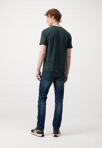 Dark green short-sleeve t-shirt, cotton material, round neck, paired with blue jeans, sitting on a relaxed fit, and black sneakers with cream accents.