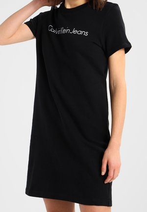 Femme portant une robe T-shirt noire Calvin Klein Jeans, debout avec un bras levé et l'autre détendu le long du corps.