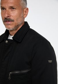 Černá bunda se strukturovaným límcem, předním zipem a kapsou na zip na hrudi; na rameni obsahuje nenápadné logo.