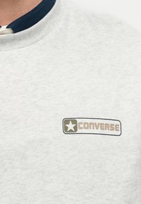 Κοντινή λήψη γκρι φούτερ με αστερίσκο σε πράσινο χρώμα και μπεζ λογότυπο "Converse" στο αριστερό στήθος, φορεμένο πάνω από πουκάμισο με σκούρο μπλε γιακά.