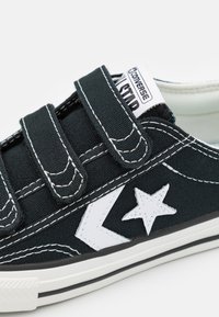Sorte lerretsneakers med justerbare borrelåser, hvite sømmer, stjerne logo og gummisåle med svart og hvite striper.
