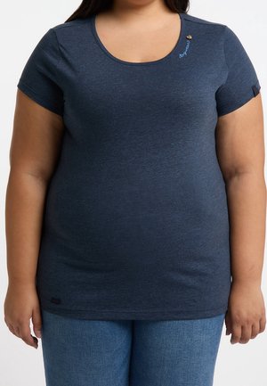 Kvinde iført en mørkeblå, tætsiddende T-shirt med korte ærmer og broderet skrift nær halsudskæringen samt mellemblå jeans.