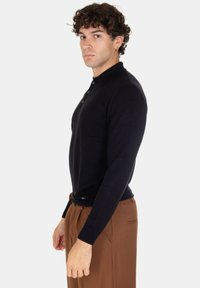 Polo shirt nera a maniche lunghe con colletto bottonato, design aderente, polsini a costine e tessuto fine, abbinata a pantaloni marroni.