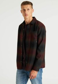 Rood en zwart geruit overhemd met knoopsluiting, enkele borstzak, gemaakt van gestructureerde stof, gedragen over een donker T-shirt, gecombineerd met lichtblauwe jeans.