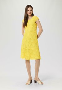 Gelbes Spitzenkleid mit kurzen Ärmeln, das ein Fächerdesign, einen gestuften Saum und ein gefüttertes Innenfutter für zusätzlichen Schutz bietet. Kombiniert mit beigen Absatzsandalen.